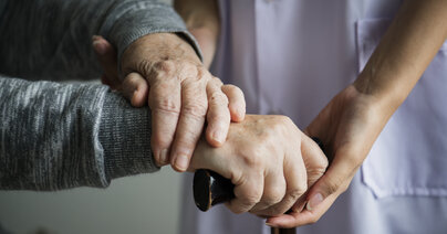zwei Paar Hände, älteres paar Hände halten Gehstock und werden von dem jüngeren Paar Händen gehalten