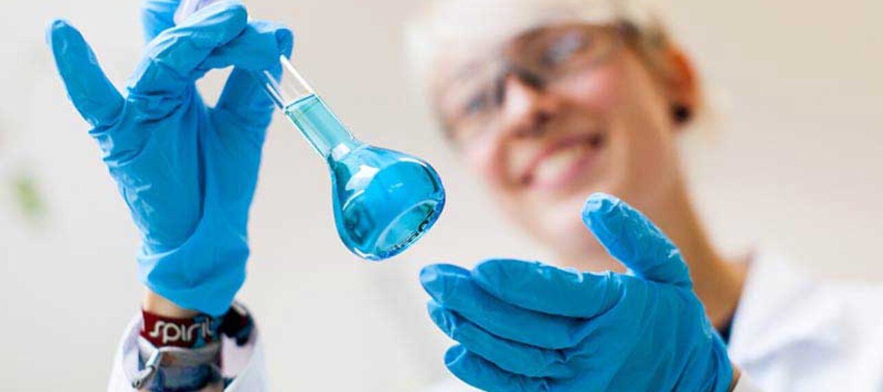 Frau mit Laborhaube und Kittel schüttelt ein Reagenzglas