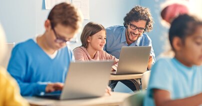 Grundschulunterricht in Informatik: Ein Mädchen benutzt Laptop, freundlicher Lehrer hilft ihr, indem er den Unterricht und die Aufgaben erklärt. Kinder erhalten moderne Bildung.
